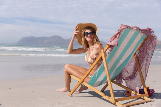Smiling caucasian woman wearing bikini sitting on deck chair looking at camera at the beach
