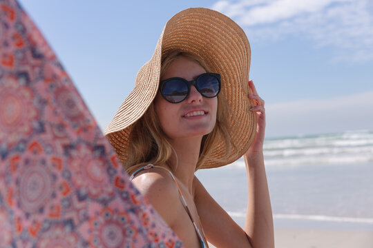 Fototapeta Smiling caucasian woman wearing bikini and hat sitting on deck chair looking at camera at the beach
