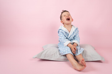 Cranky child boy refuses to go to bed. Kid boy in bathrobe sits on pillow on pink background