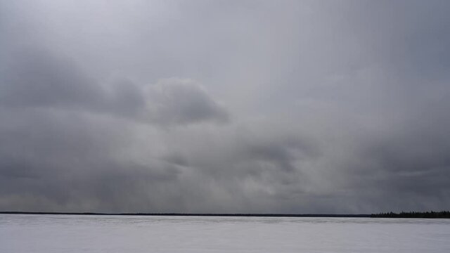 Grey storm clouds move above a frozen lake that is covered by snow. A thin line of trees can be seen on the horizon.
