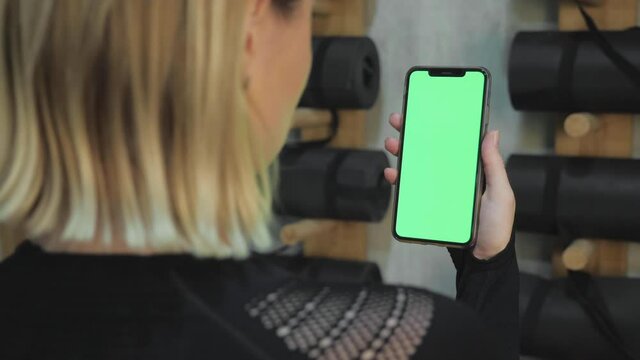 back view of fitness woman using green screen smart phone at home. Woman preparing for workout using smart phone. Woman doing a tap on mobile phone while resting during fitness training