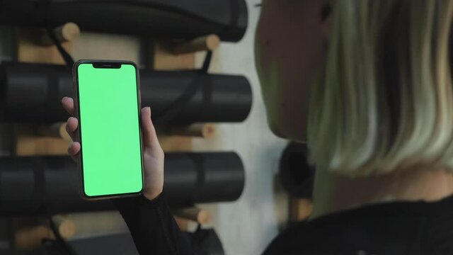 A Young Athletic Woman Holds A Smartphone in Her Hand, Shows A Green Chromakey Screen, Is in The Gym