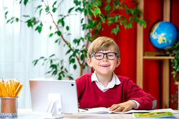 Smart happy preteen boy boy with eye blue light blocking glasses while using computer laptop and notebook studying online lessons. Work at home remotely doing homework. Kid in uniform