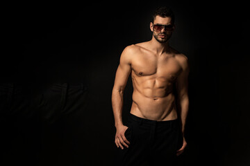 Fototapeta premium Muscle strong beautiful stripped male model in black trousers wearing glasses eyewear on black isolated font background
