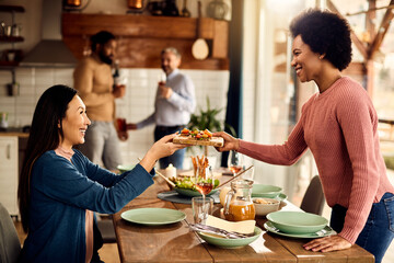 Happy African American woman serving appetizer while setting table for lunch with her friends at home.