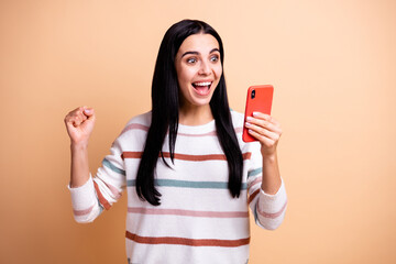 Photo of young attractive charming happy excited crazy girl hold fist in victory hold phone isolated on beige color background