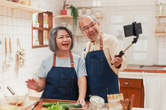 Portrait of mature man and woman pensioner using smartphone taking photo selfie together in the kitchen at home. Cheerful Asian senior couple having fun cooking healthy food together.