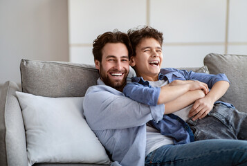 Fatherhood concept. Cheerful dad embracing son, tickling and laughing while resting on couch at...