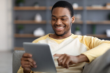 Positive black man reclining on sofa, using digital tablet, closeup