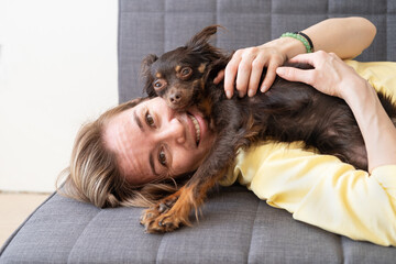 Happy blonde woman lying wth funny brown russian toy terrier on couch