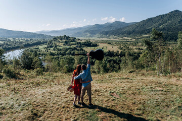 A couple of travelers looking at the Carpathians with a guitar in their hands, a river flows at the bottom of the mountains. Morning in the mountains.Travel and leisure. 