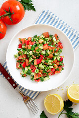 Vegetable salad in a white plate on a light background top view. Salad with tomatoes, cucumbers, peppers, parsley and lemon. Vegan or diet food.