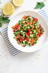 Vegetable salad on a white plate on a light background. Salad with tomatoes, cucumbers, peppers, parsley and lemon. Vegan or diet food. Copy space for text.