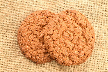 Two dark brown oatmeal cookies on burlap, close-up, top view.