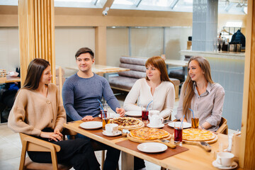 A group of young cheerful friends is sitting in a cafe talking and eating pizza. Lunch at the pizzeria.