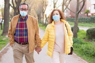 Senior caucasian couple walking in the park holding hands. Both are wearing a protective mask....