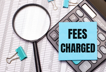 On the table are reports, a magnifying glass, a calculator, and a blue note sticker with the words FEES CHARGED. Business concept
