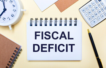 On a light background, a white alarm clock, a calculator, a pen and a notebook with the text FISCAL DEFICIT. Flat lay