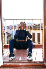 Naklejka na ściany i meble black afro woman dressed in sportswear listening to music on headphones very happy on the balcony because she is going to start exercising at home due to the covid19 coronavirus pandemic