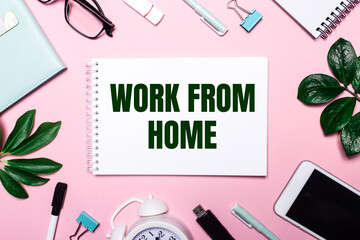 Fototapeta na wymiar WORK FROM HOME is written in a white notebook on a pink background surrounded by business accessories and green leaves.