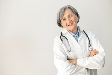 Positive senior mature female doctor with a stethoscope in a medical coat posing isolated over gray background, standing with arms folded, smiling and welcoming healthcare worker looking at the camera