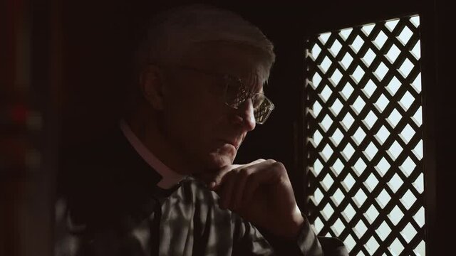 Lockdown of elderly Caucasian priest wearing robe, white collar and glasses sitting in confessional with Bible in hands and having conversation with parishioner through wooden window