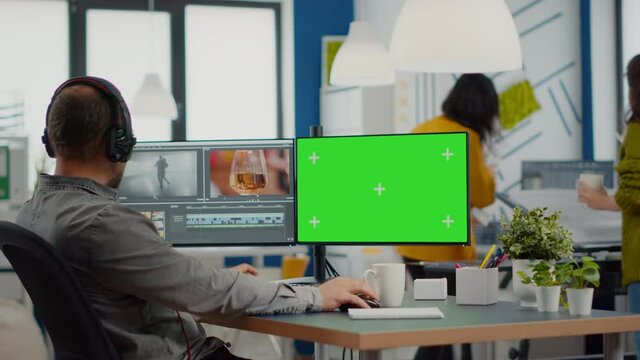 Videographer using computer with chroma key, mock up isolated display editing video and audio footage. Man editor processing film montage on pc with green screen while team working in background