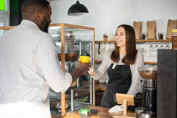 Friendly and helpful waitress is serving an African-American male customer, preparing and giving paper cup of coffee to multiracial guy. Woman in apron holds out take-away coffee to guest of cafe