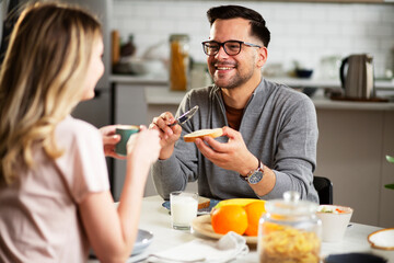 Beautiful man enjoying in breakfast with his girffriend. Happy young couple drinking coffee and eating sandwich at home.