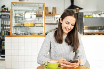 Smiling young brunette woman enjoys a morning coffee sitting in the cozy cafe and chatting on the smartphone, female student has a break with phone and hot drink in the cafeteria