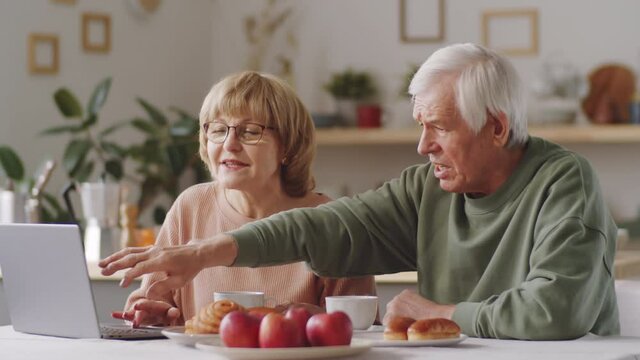 Senior man pointing on laptop screen, talking with cheerful wife and embracing her while sitting together at kitchen table and having tea