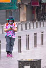 Asian female tourist using smartphone while standing between row of metal barriers during walking down on the street in vertical frame