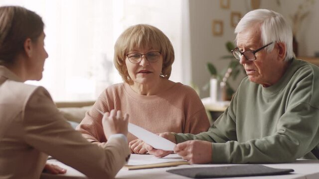 Elderly man and woman sitting at table, reading contract and discussing details with female insurance agent during consultation at home