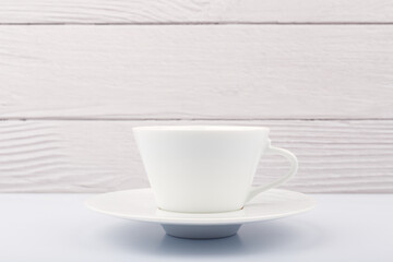 Close up of white ceramic coffee or tea cup with saucer on white table against white blurred wooden background with copy space