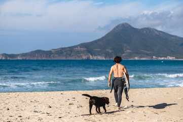 rear view shirtless man with diving suit holding a surfboard walking on a sandy beach with his best friend a chocolate labrador.