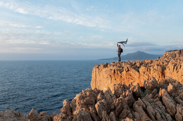 Young man standing on a high jagged cliff waving a scarf. Looking the sunset on the sea