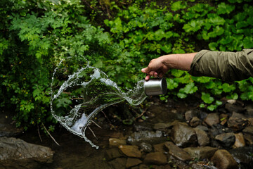 Naklejka premium A splash of fresh water from a mug in the hands of a man on the background of nature. Drinking water concept. A splash of freshness. Natural water. Healthy lifestyle. cold water from a mountain river