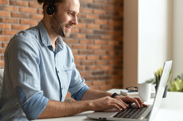 A confident male office worker sitting at the desk with a laptop, using headset communicating with...