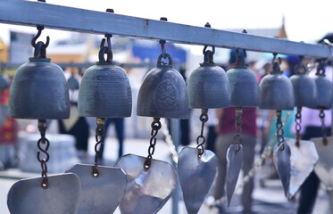 A row of Thai bronze bells hanging from a metal bar at the Thai temple, with people and buildings...