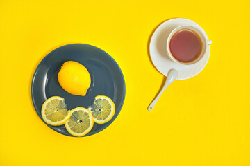 fresh lemon whole and sliced on plate. there is cup of tea with saucer and spoon on yellow table