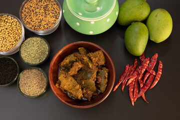 Mango pickle or Traditional spicy Indian mango pickle chillies and spices with ceramaic jar and raw mango, selective focus top view isolated on black