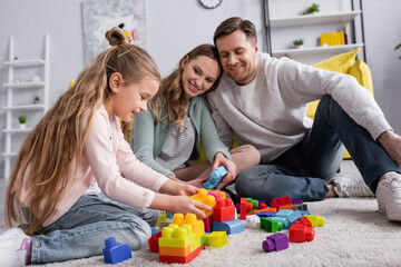 Smiling kid playing building blocks on carpet near blurred parents.