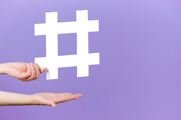 Concept of trendy social media posts and blogging. Female hands holding large big white hashtag sign, viral web content, internet promotion, isolated on purple studio background with copy space