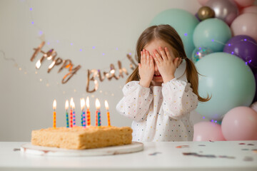 cute little girl blows out candles on a birthday cake at home against a backdrop of balloons. Child's birthday