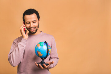 Cheerful african american black man holding the globe with love and care isolated over beige background. Travel concept, looking for a jorney. Using mobile phone.