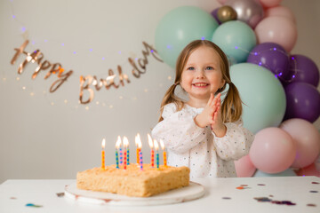 cute little girl blows out candles on a birthday cake at home against a backdrop of balloons....