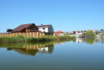 Naklejka na ściany i meble Modern cottage houses reflected in the water built on the bank of the lake or river. Constructing houses along the river with a beautiful view on the river.