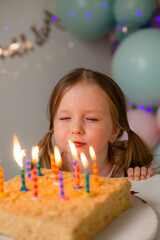cute little girl blows out candles on a birthday cake at home against a backdrop of balloons....