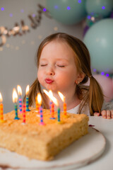 cute little girl blows out candles on a birthday cake at home against a backdrop of balloons....