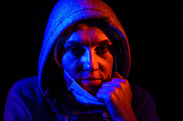 Portrait of a young woman in a hood, wearing a protective medical mask, illuminated by red and blue light, isolated on black background